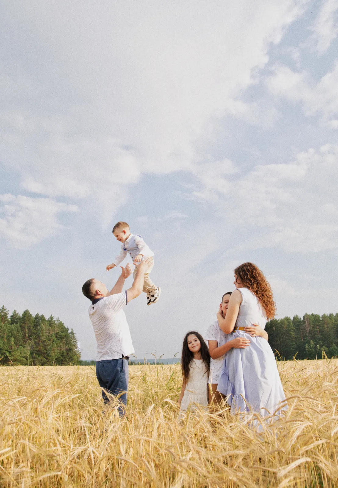Father tossing his laughing toddler into the air in a wheat field while the rest of the family looks on