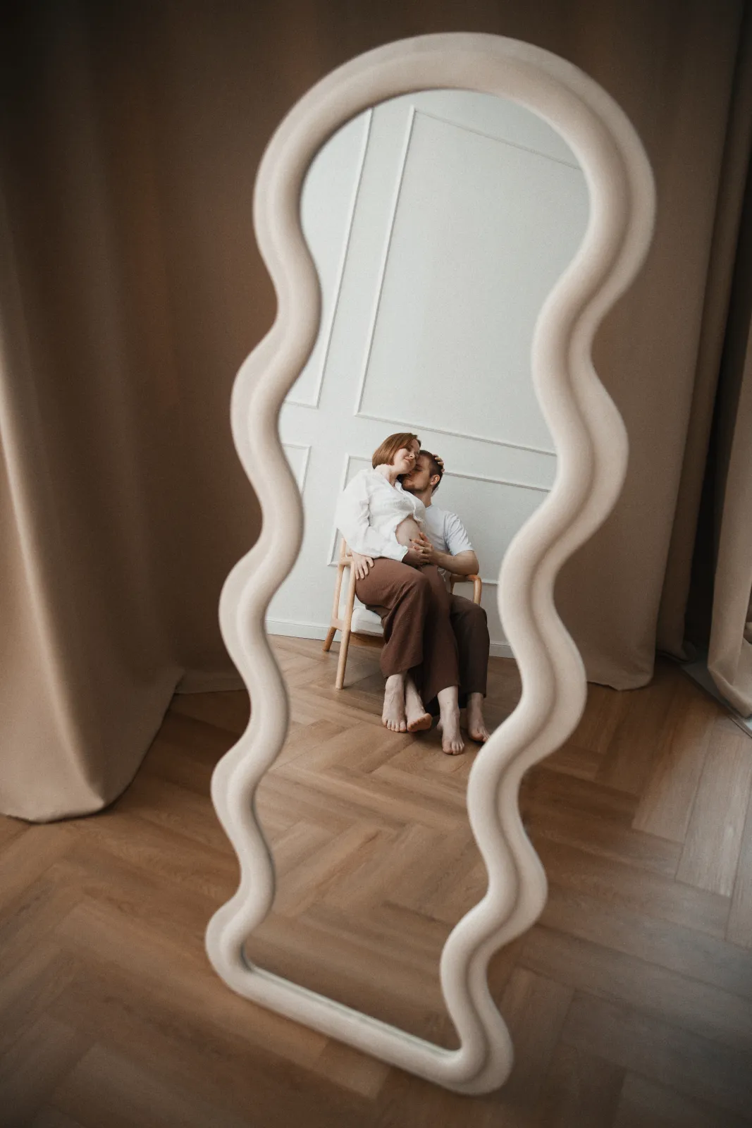 Expecting couple embracing on a chair, seen through the reflection of a wavy full-length mirror