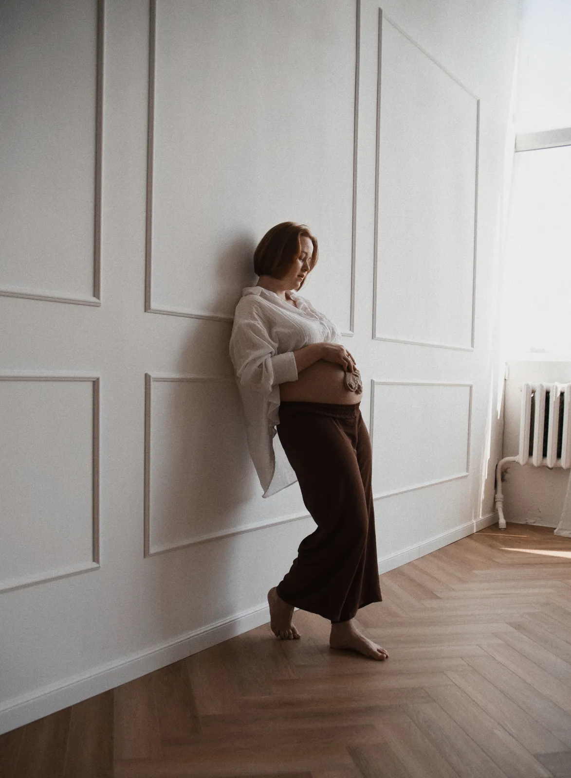 Pregnant woman leaning against a paneled wall, holding tiny baby booties on her bump