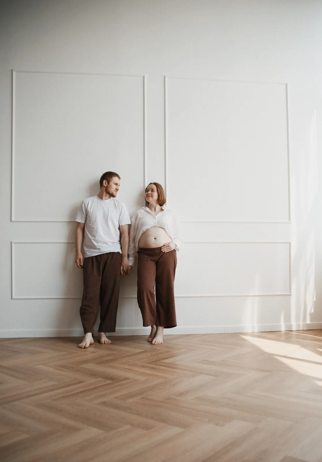 Expecting couple leaning against a white paneled wall, holding hands and looking at each other